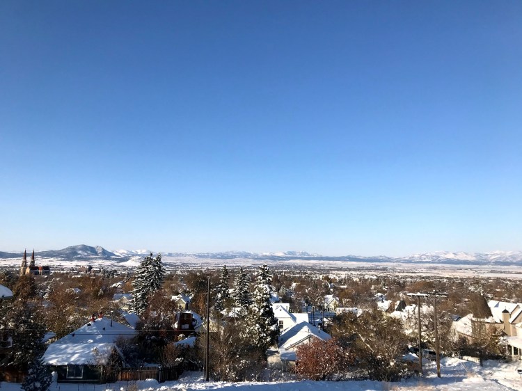 Snow covered town as viewed from a tall hill overlooking a wide valley with other mountains in the distance.