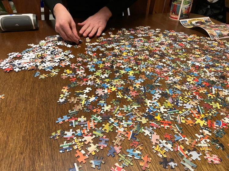 Wooden table covered in multi-colored puzzle pieces that have not been put together. In the background a man's hands are sorting the pieces.