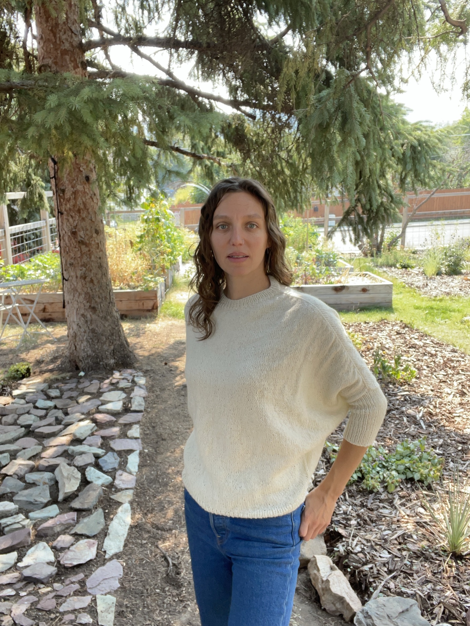 A white woman with wavy brown hair wearing an off-white sweater and blue jeans stands in a garden