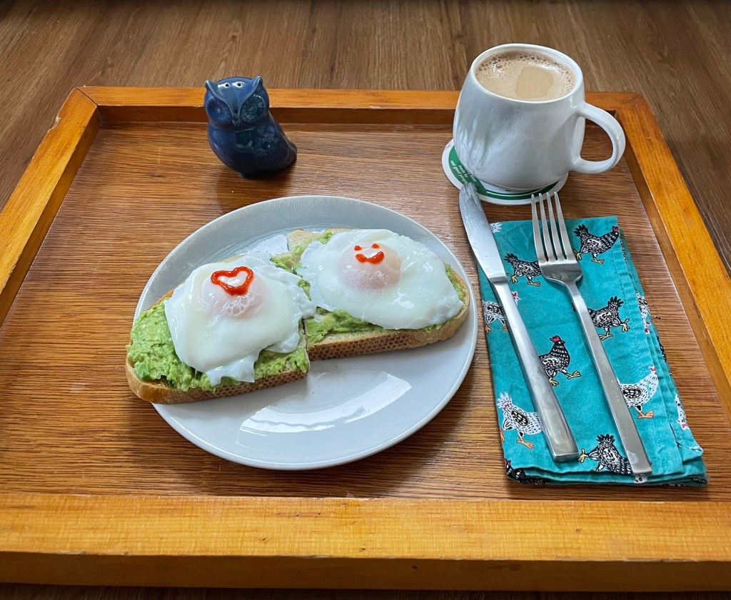 Wooden breakfast tray with creamed coffee, plate of two poached eggs on avocado toast, a blue owl salt shaker, and fork and knife on a cloth napkin with chickens on it. The yolks of the poached eggs have a heart and a smiley face drawn on them with red sriracha sauce.