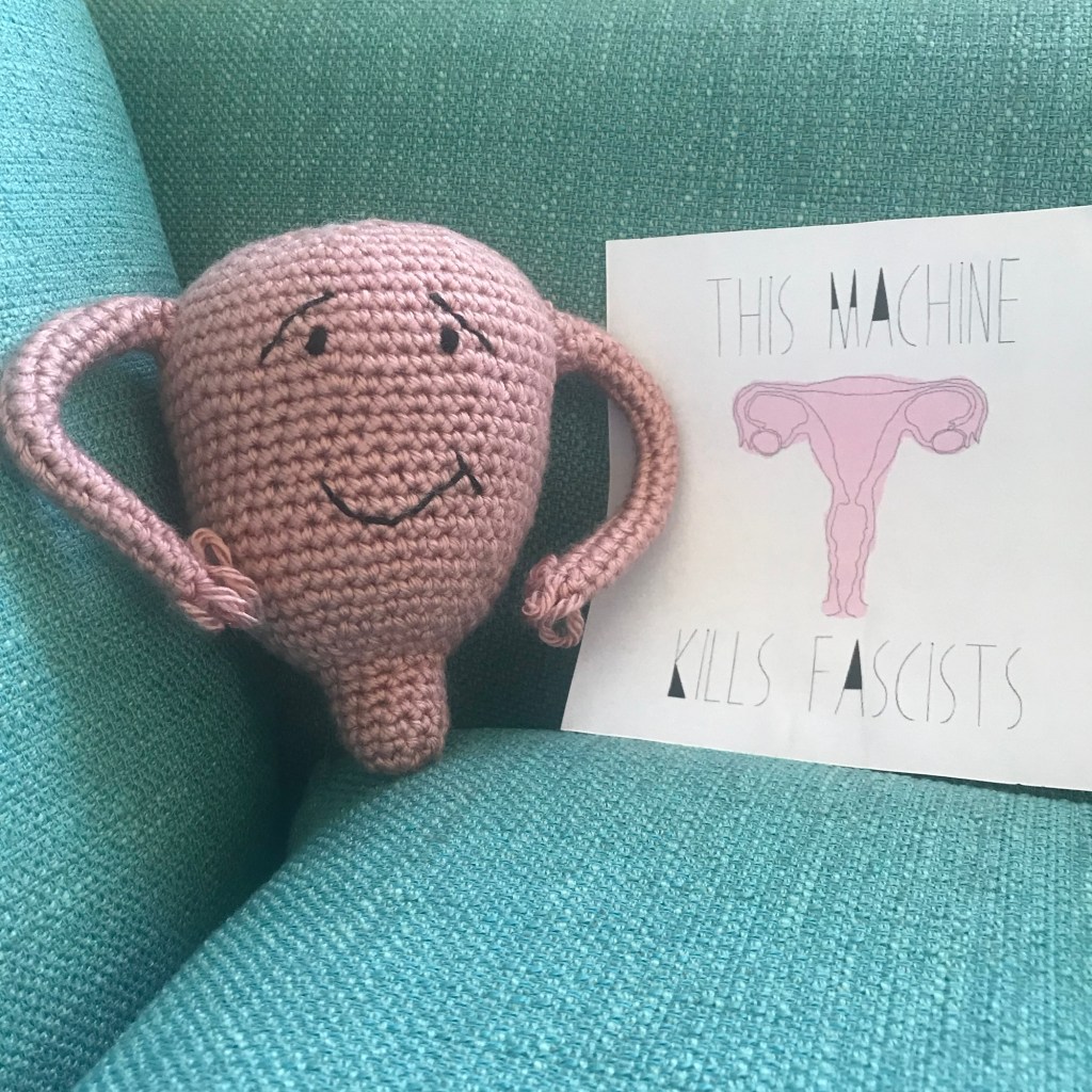 Pink, crocheted uterus with an embroidered smirking face, on a teal couch to the left of an image of a uterus with the slogan “this machine kills fascists” surrounding it.