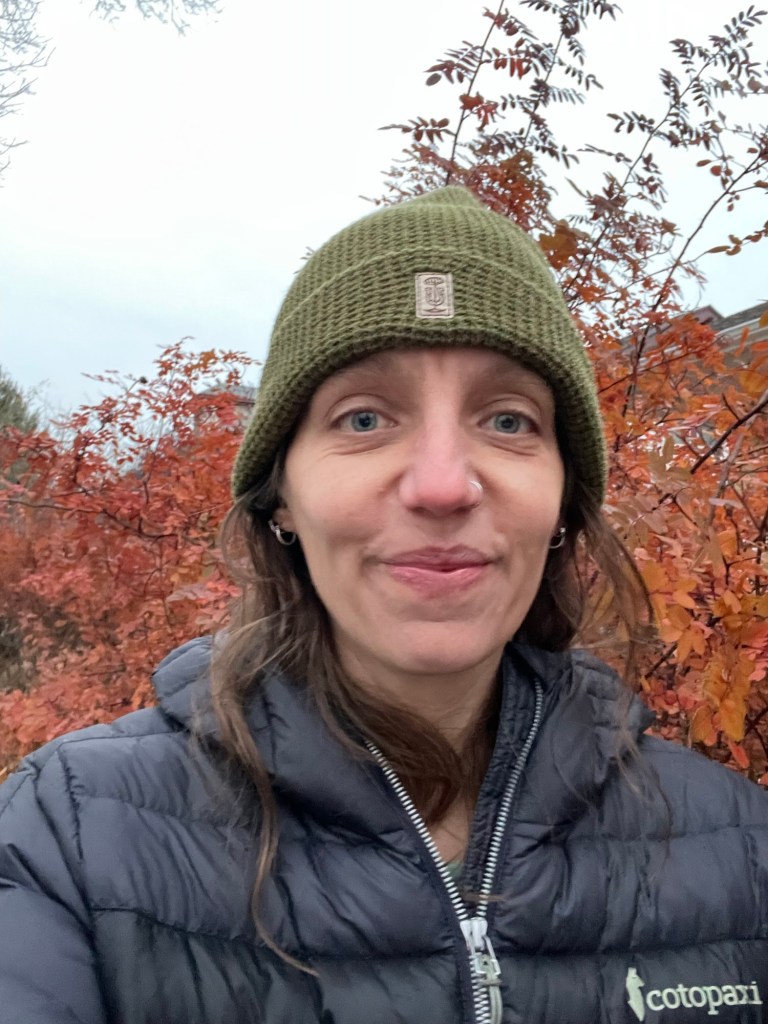 White woman in olive drab, knitted hat with black puffer jacket in front of a bush with small, compound, reddish-orange leaves